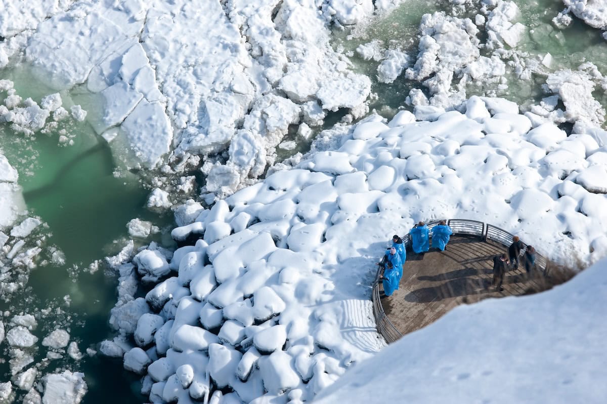 Menschen stehen auf einer Aussichtsplattform über dem gefrorenen Niagara mit zerbrochenem Eis und schneebedeckten Felsen. Luftaufnahme einer Winterszene an den Niagarafällen, die kaltes Wetter, Eisschollen und einen Aussichtspunkt für Touristen zeigt.