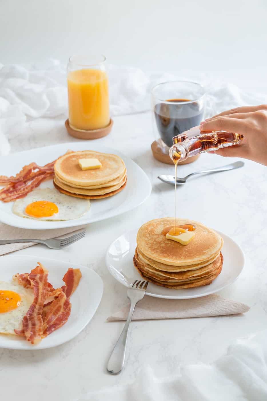 Frühstück mit Pancakes, Spiegelei, Bacon, Orangensaft und Kaffee. Auf die Pancakes wird Ahornsirup geschüttet.