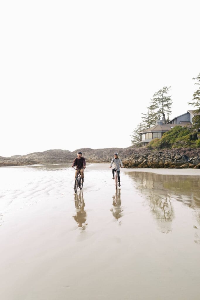 Ein Paar fährt mit dem Fahrrad entlang des North Chesterman Beach in Tofino. Das kanadische Hotel Wickaninnish Inn ist im Hintergrund.