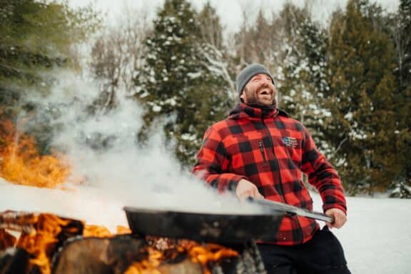 Ein Mann steht lachend am Feuer und bereitet Essen in einer großen Pfanne zu.