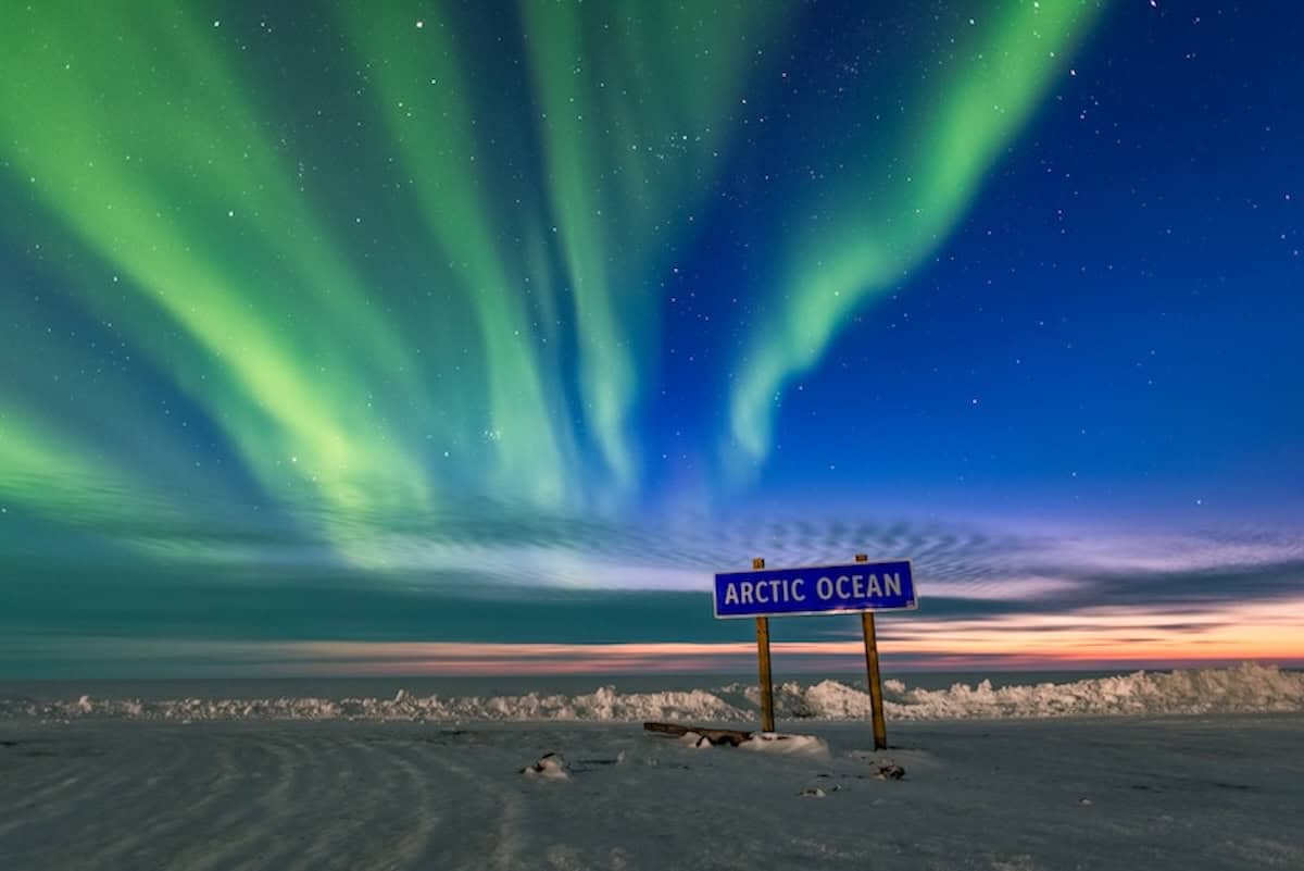 Schild mit der Aufschrift "Arctic Ocean" steht vor dem Meer bei Nacht im Schnee. Darüber sind Polarlichter zu sehen.